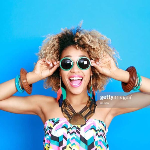 Summer portrait of happy afro american young woman wearing summer clothes and sunglesses, standing against blue background and laughing at camera.